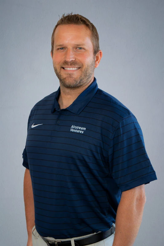 Man in blue polo shirt smiling, standing against gray background.