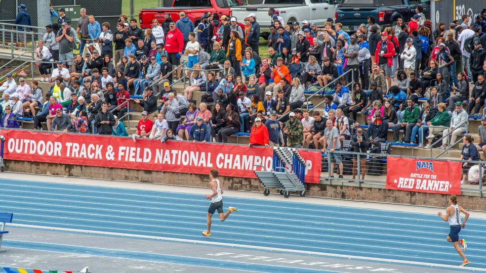 Fast runners competing in a track race at an outdoor stadium with a large crowd watching.