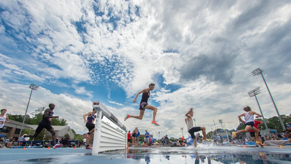 Jumping athlete clearing hurdle at outdoor track meet under cloudy sky.